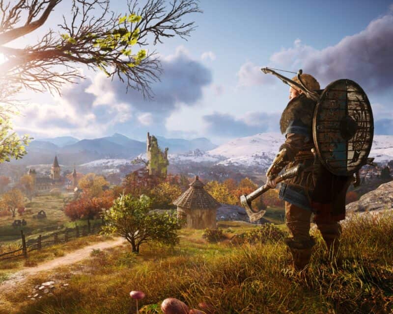 Zrzut ekranu z gry Assassin’s Creed Valhalla. Przedstawia on blondwłosego mężczyznę - wikinga, ubranego w strój do walki, z tarczą i łukiem na plecach oraz toporem w ręce. Stoi on na pagórku i patrzy w dół na wioskę. Prawdopodobnie trwa jesień, kolorystyka jest ciepła - pomarańczowo-zielona. Po prawej stronie od wioski widnieją przykryte śniegiem góry.