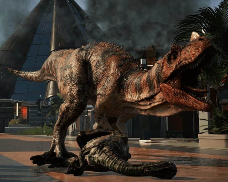 Zapowiedź Jurassic World Evolution 2 - na obrazku Tyranozaur tryumfuje nad pokonanym zauropodem. W tle widać alejki głównego placu w parku.