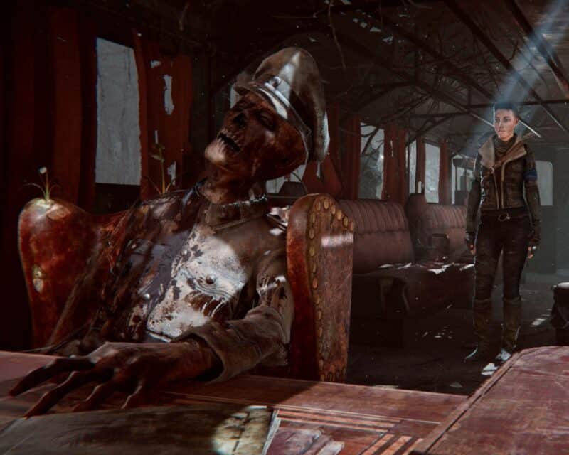 Kobieta z krótkimi włosami i w skórzanej kurtce stoi w zrujnowanym pociągu, w ruinie wagonu znajduje się biurko, a za nim siedzi trup z resztkami czaszki, w stroju nazisty, wokół mnóstwo rupieci i ogólny bałagan, Syberia The World Before data premiery
