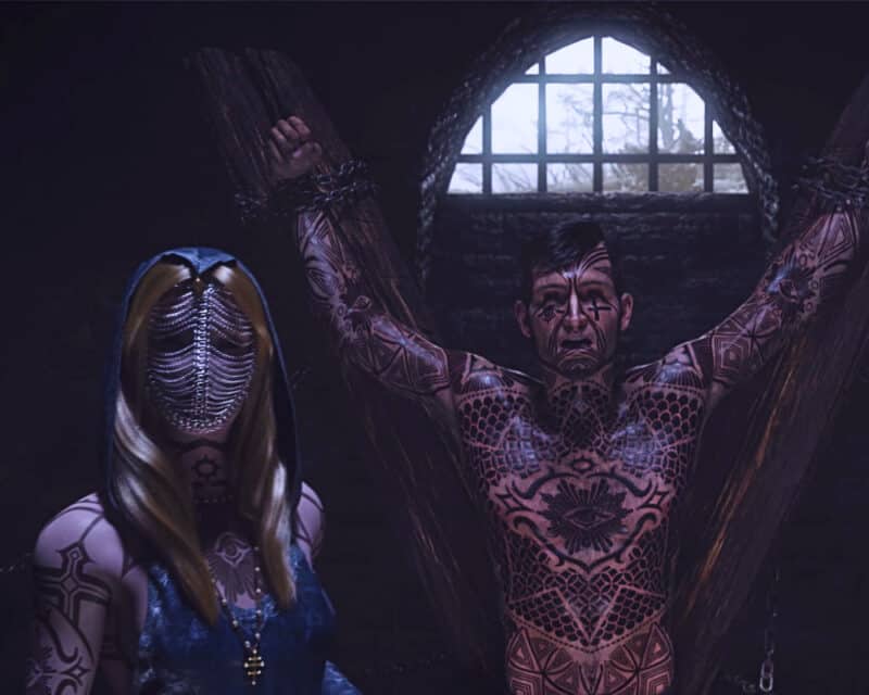 Disciples: Liberation znamy wymagania sprzętowe gry - na obrazku wytatuowany mężczyzna jest uwięziony w lochu, jego oprawca to elegancka kobieta o zasłoniętej twarzy.