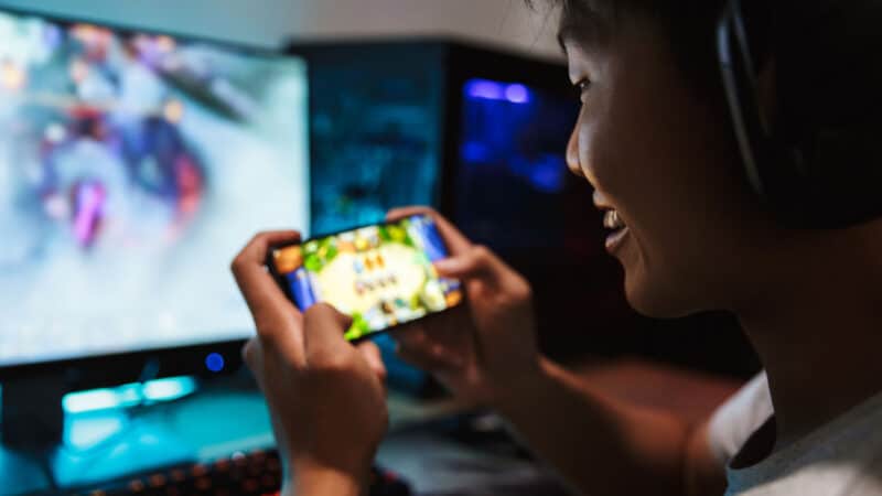 Azjata grający na urządzeniu mobilnym, w tle monitor z włączoną grą.