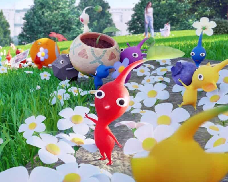 Pikmin Bloom dostępny w Polsce - na obrazku uśmiechnięte Pikminy niosą rzeczy po ukwieconej parkowej alejce. W tle kobieta spaceruje z psem.