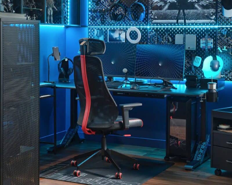 meble IKEA dla graczy, na zdjęciu ciemno, niebieska poświata, na środku czarno-czerwony fotel przysunięty do biurka, na którym stoi monitor, wokół mnóstwo akcesoriów