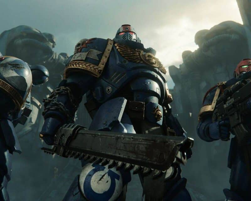 Grafika z gry Warhammer 40,000: Space Marine 2, przedstawia trzech Marines w ich charakterystycznych błękitnych ciężkich zbrojach, stoją przodem do kamery, jeden z nich trzyma w prawej dłoni piłomiecz