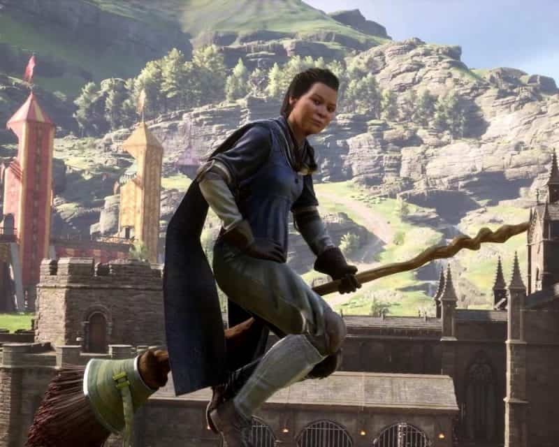 kobieta w zielonym płaszczu, krótko obcięte ciemne włosy, siedzi na miotle w powietrzu, w dali mury zamku i piasek, Hogwarts Legacy