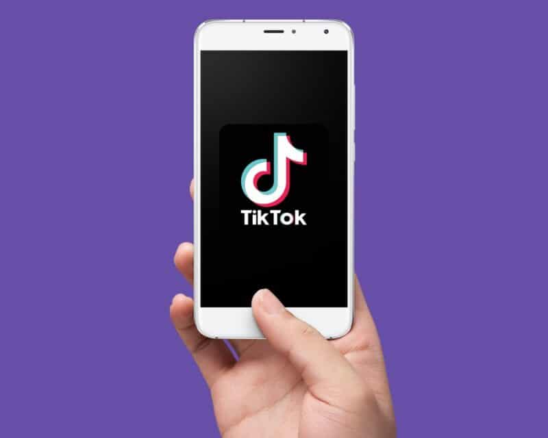 fioletowe tło, na nim ręka trzymająca telefon z czarnym ekranem, na ekranie logo TikTok