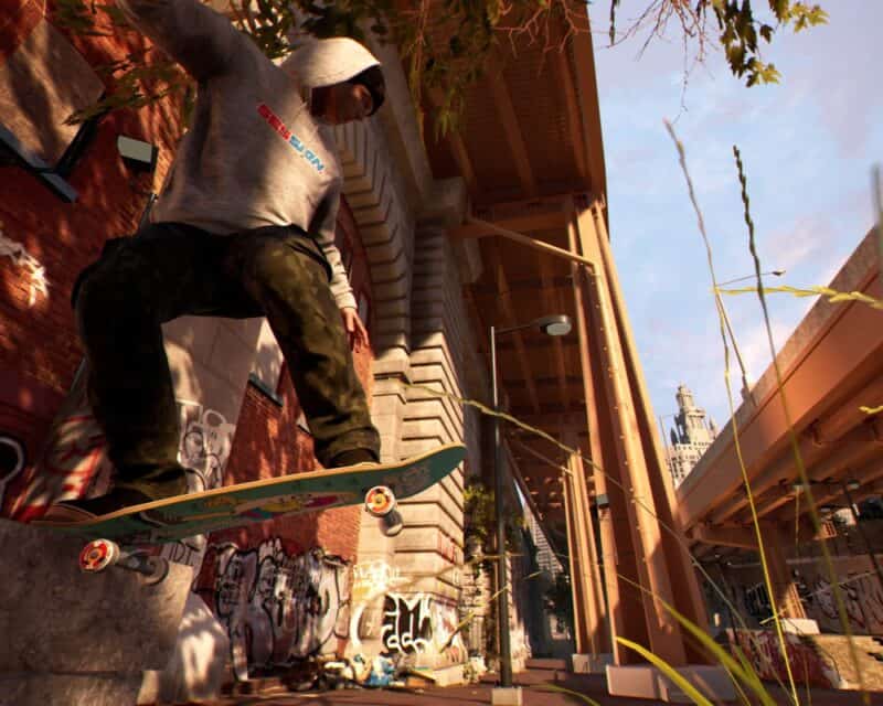 obrazek przedstawiający ubranego w czarne dresy i szarą bluzę z kapturem mężczyznę z deskorolką podczas wykonywania skoku z podwyższenia na asfalt, dookoła mury i ściany pomalowane graffiti, w tle most, Session Skate Sim data premiery