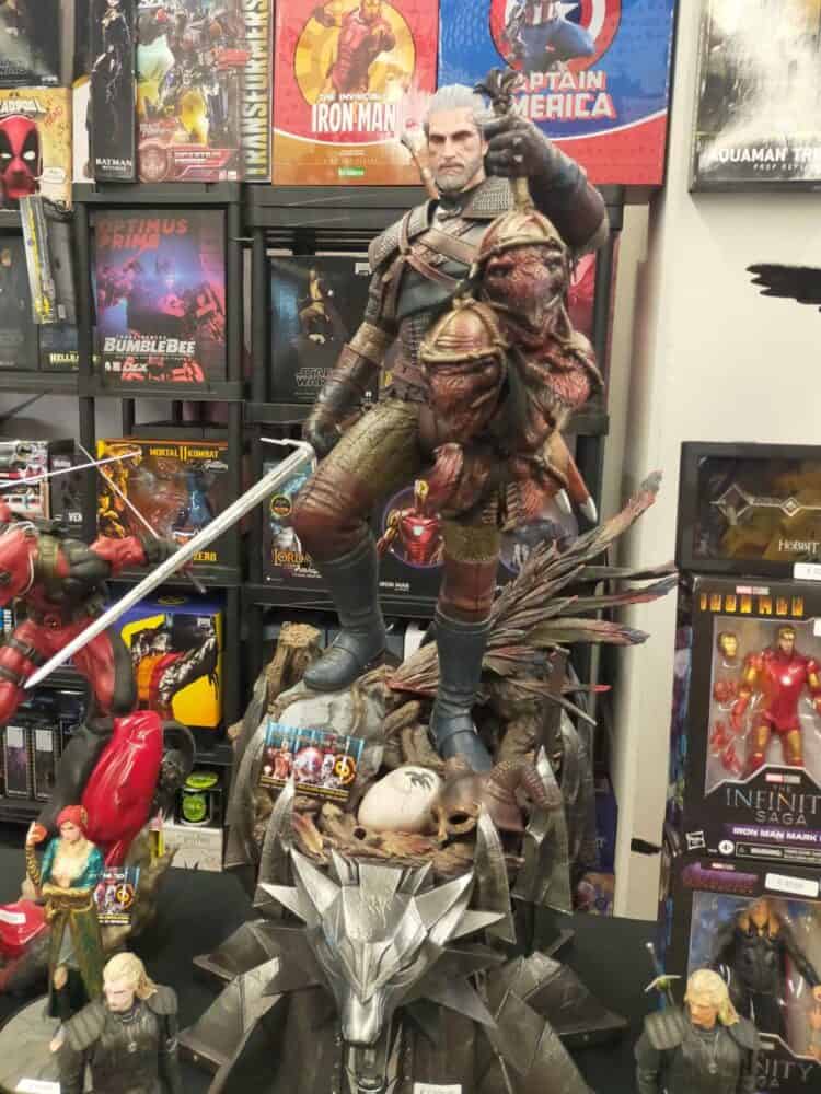 zdjęcie przedstawia figurkę Geralta, który w swojej prawej dłoni trzyma miecz, a w lewej unosi do góry odcięte głowy bestii, stoi na jakichś czaszkach i hełmach, a poniżej widać logo szkoły Wilka, za figurką widać dużo innych przedmiotów na sprzedaż