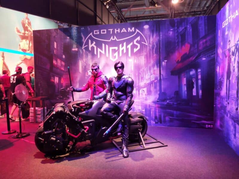 zdjęcie przedstawia dwóch mężczyzn, którzy przebrani są w stroje podobne do stroju Batmana, siedzą oni na motocyklu na tle baneru promującego grę Gotham Knights, relacja gamescom 2022
