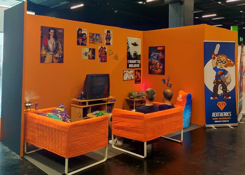 zdjęcie przedstawia kącik stylizowany na salony retro, na ścianach wiszą plakaty i nalepki, m.in. młodego DiCaprio czy starych gier, widać też stary telewizor i dwie pomarańczowe kanapy, na jednej z nich siedzi dwóch mężczyzn