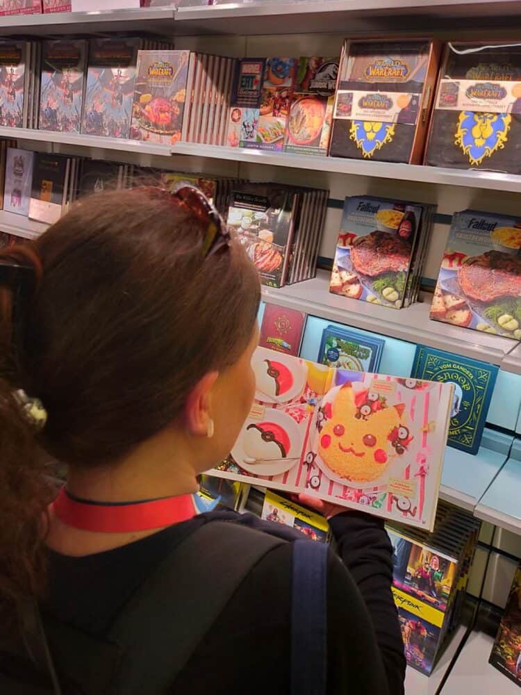 zdjęcie przedstawia kobietę (perspektywa jakbyśmy czytali jej przez ramię), czytającą książkę kucharską w klimatach Pokemonów. Na otwartej przez nią stronie widać ciastko stylizowane na Pikachu, a obok niego mniejsze, przypominające Pokeballe, w tle widać dużo innych książek, relacja gamescom 2022