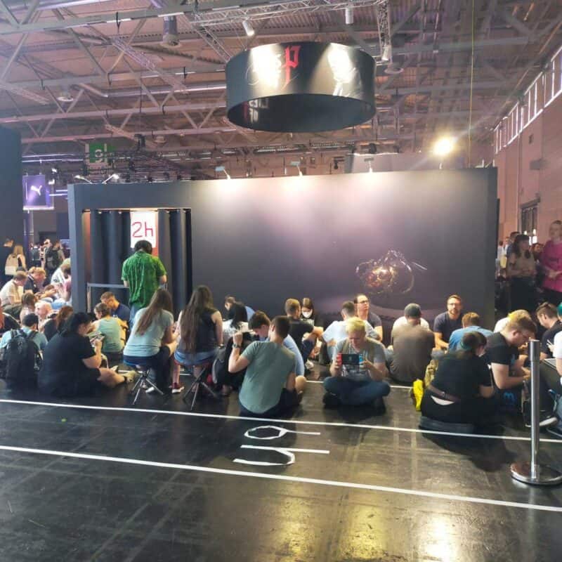 zdjęcie przedstawia kawałek stoiska Lies of P, widać czekających na możliwość zagrania ludzi, część stoi, większość siedzi na podłodze. Bliżej widać białą taśmę i napis "6h"