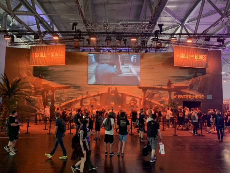 zdjęcie pochodzi ze strony jaxon.gg i przedstawia fragment dekoracji dla gry Skull and Bones, ścianka jest stylizowana na statek piracki, na górze powieszony jest telewizor, po prawej znajduje się wejście, widać także czekających w kolejce do zagrania ludzi