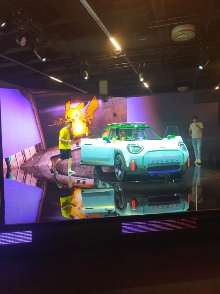 zdjęcie przedstawia samochód Cooper Mini, ten w kolaboracji z Pokemonami, z otwartymi drzwiami, siedzą w nim dwie osoby. Z tyłu na ścianie widać Pikachu, a obok samochodu kręcą się dwie osoby