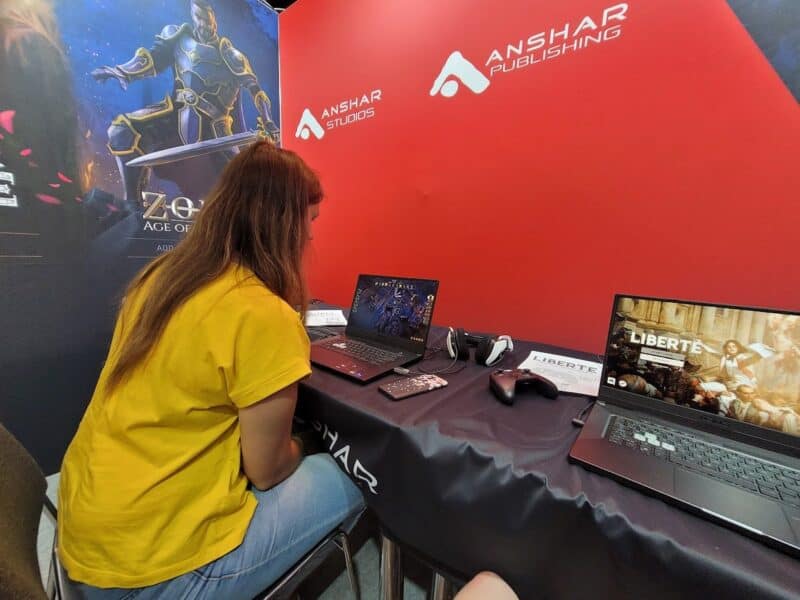 zdjęcie przedstawia kobietę w żółtej bluzce grającą w coś na laptopie, obok inny laptop z odpaloną inną grą, laptopy leżą na czarnym materiale a ścianka za stolikiem ma czerwony kolor