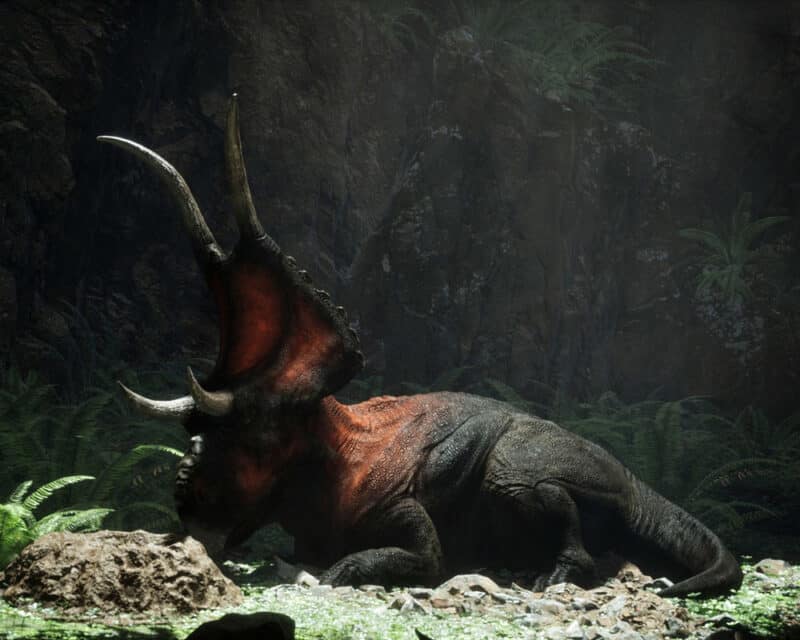 Grafika przedstawia dinozaura z gry Instinction. Gad o umaszczeniu czarnym z domieszką czerwieni, leży spokojnie pod skalną ścianą.