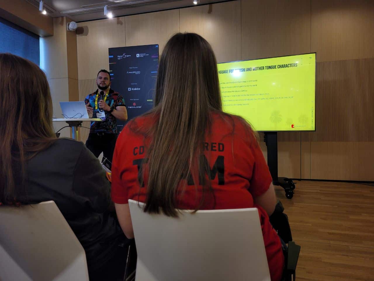 Digital Dragons 2023, o lokalizacji Cyberpunka, widać ekran na drugim planie, na pierwszym siedząca przede mną słuchająca, obok niej, na drugim planie, prowadzący