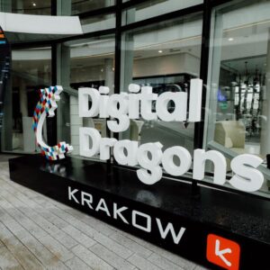 zdjęcie loga konferencji Digital Dragons w formie 3D stojącego przed centrum konferencyjnym, logo ma nazwę konferencji zapisaną białą czcionką i pod spodem na czarnym tle napis KRAKÓW. W tle przeszklone szyby