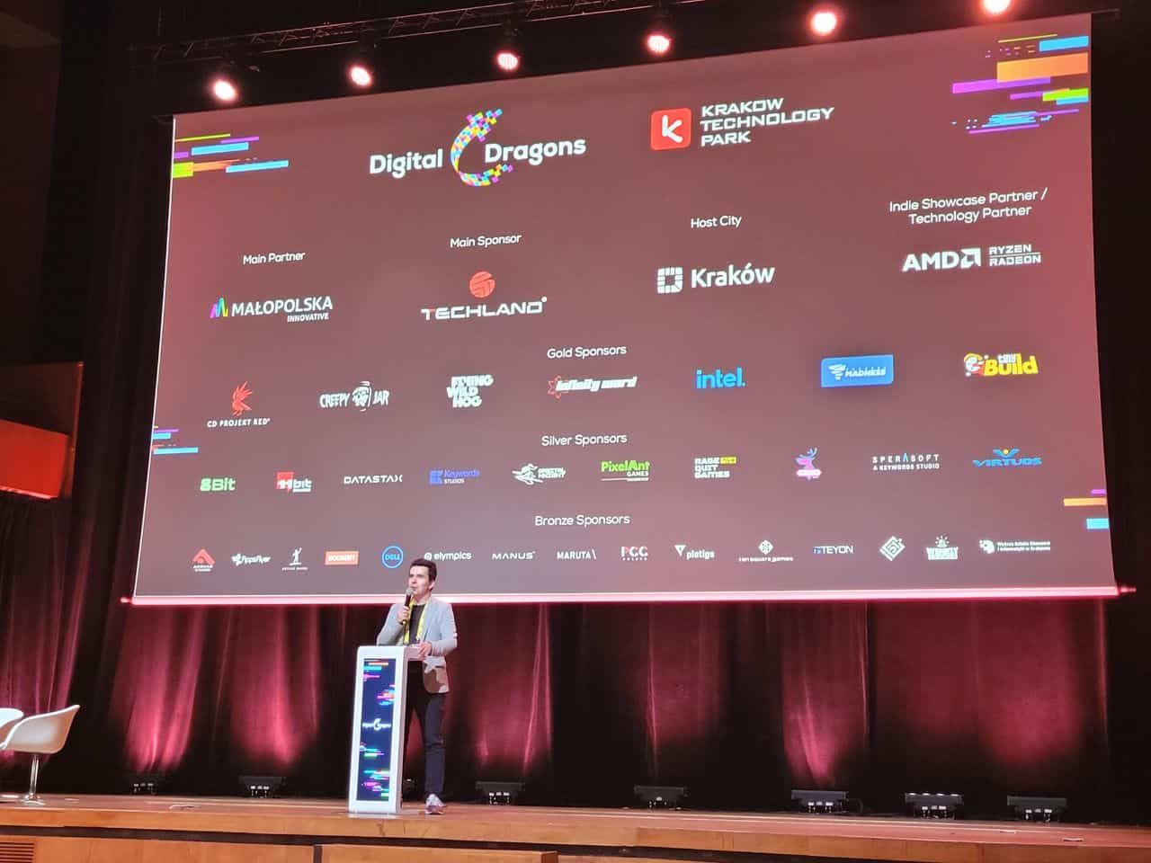 Digital Dragons 2023, zapowiedź kolejnego wykladu, widzimy mężczyznę stojącego na scenie przy podium, a za nim ekran z logami wszystkich partnerów wydarzenia