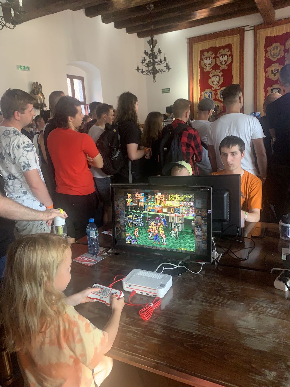 Na obrazku widać dziewczynkę, która gra w retro gierkę na konsoli oraz tłum ludzi za nią. 
