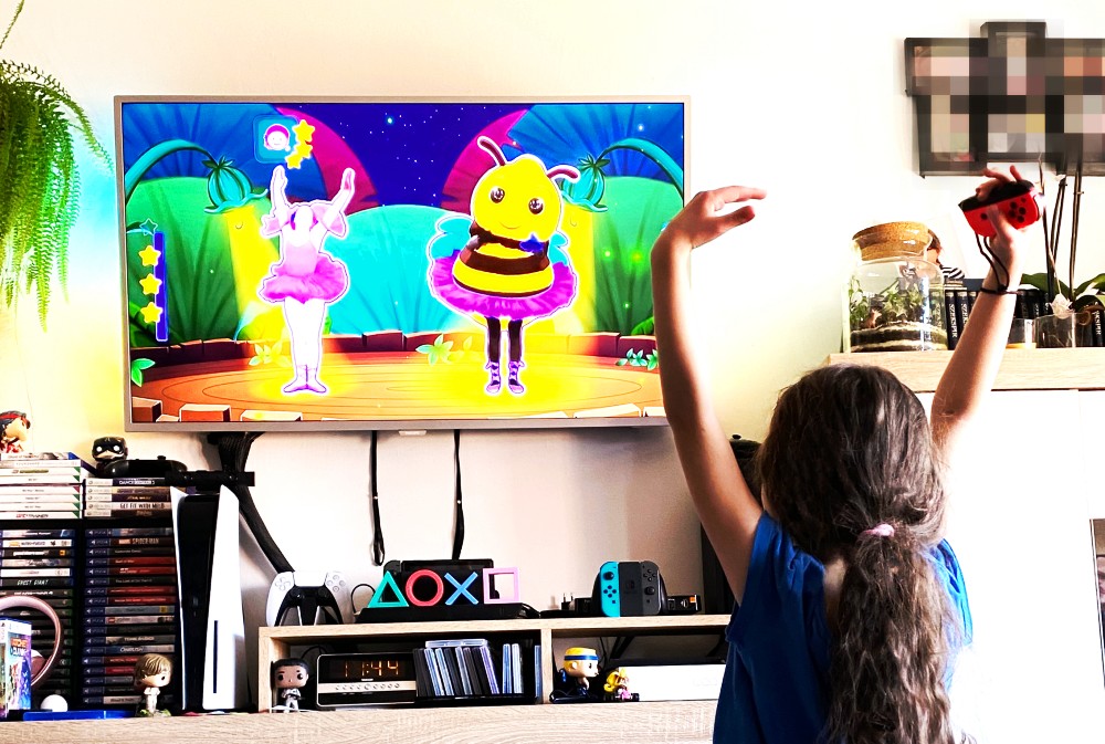 Dziewczynka grająca w grę wideo Just Dance. Widzimy telewizor z podglądem gry oraz tańczącą dziewczynkę.