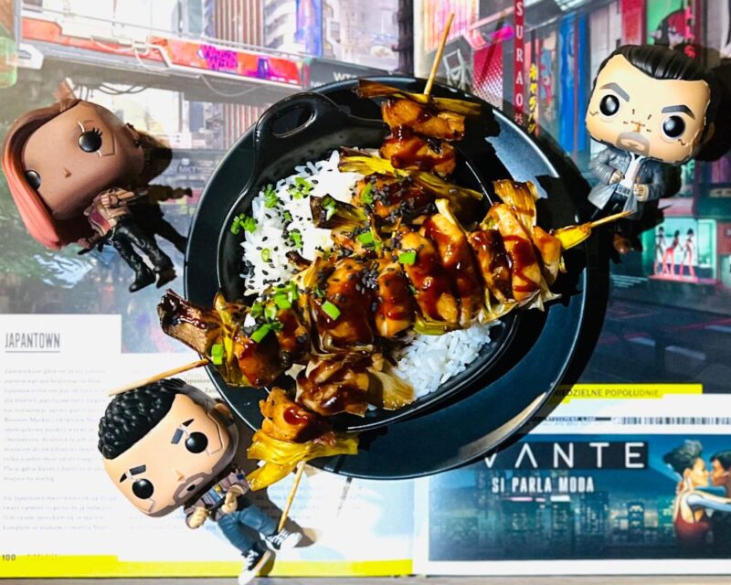przepis na cyberpunkowe yakitori z gry cyberpunk 2077, na zdjęciu widać talerz z dwoma patyczkami z nadzianymi na nie kawałkami mięsa, wokół talerza leżą figurki Funko przedstawiające postaci z gry