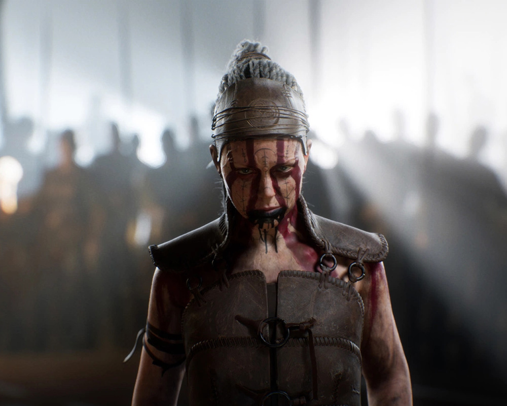 Protagonistka gry Senua's Saga: Hellblade 2 – Senua. Kobieta stoi samotnie na pierwszym planie. Ma przygnębiony wyraz twarzy (pokrytej tatuażami), która spływa krwią. Ma na sobie skórzany stroj do walki i odkryte ramiona. Włosy spięte w kucyk. Za nią widnieje rozmazana grupa ludzi, prawdopodobnie szykujących się do walki.