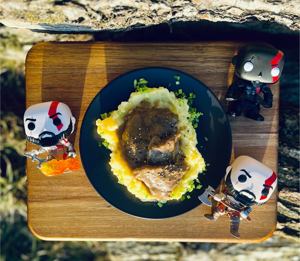 Przepis na bitki z minotaura, na zdjęciu widzimy rzut z góry na deskę z gotową potrawą, po jej bokach leżą 3 figurki Kratosa