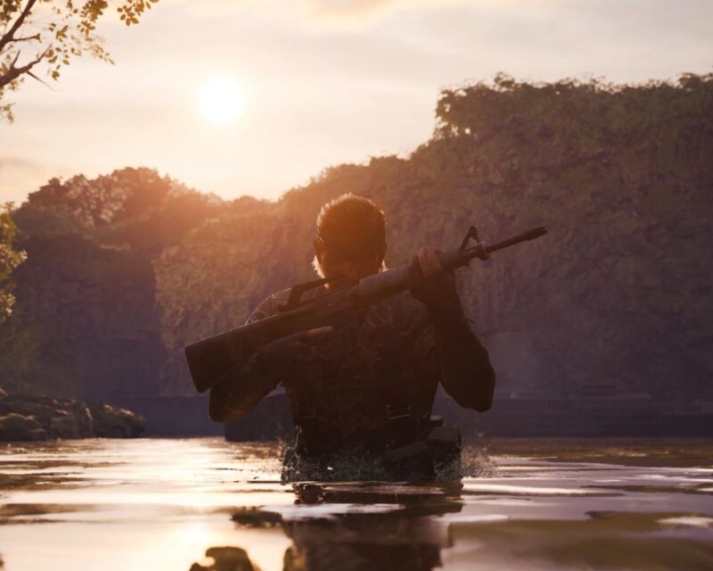 Główny bohater gry stoi do pasa w rzece. Trzyma karabin. W tle widać góry i zachodzące słońce.