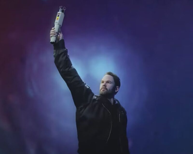 Alan Wake 2: Night Springs, widzimy młodego mężczyznę z zarostem i krótkimi włosami, ubranego w kurtkę. Trzyma dziwny sprzęt w ręce wyciągniętej do góry, w tle głęboki ciemny fiolet i na środku za postacią w oddali widać światła