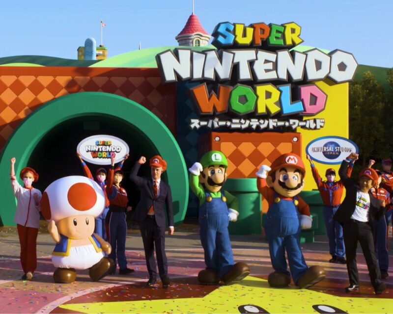 Grupa ludzi stoi na przeciwko japońskiego parku rozrywki Super Nintendo World. Machają do kamery. Niektórzy są przebrani za postaci z gier Mario.