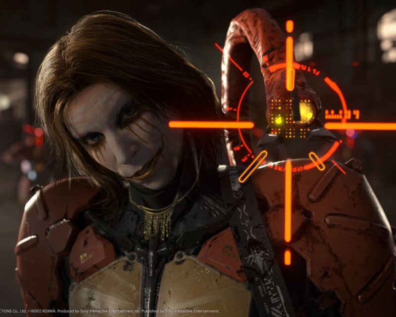 Na obrazku znajduje sie postąc z gry death stranding. Jest to brunet z włosami do ramion. Na twarzy ma makijaż przypominający ten z zespołu KISS. Nosi pomarańczową, metalową zbroję, za której wyłania się metaliczny wąż lub macka. Ewidetnie celuje on w nas lub skanuje.