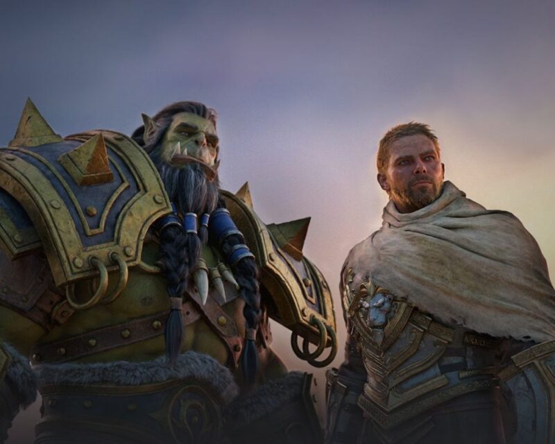 Wielki, zielony ork stoi obok ubranego w zbroję człowieka. Uśmiechnięci patrzą gdzieś przed siebie. Za ich plecami zachód słońca.