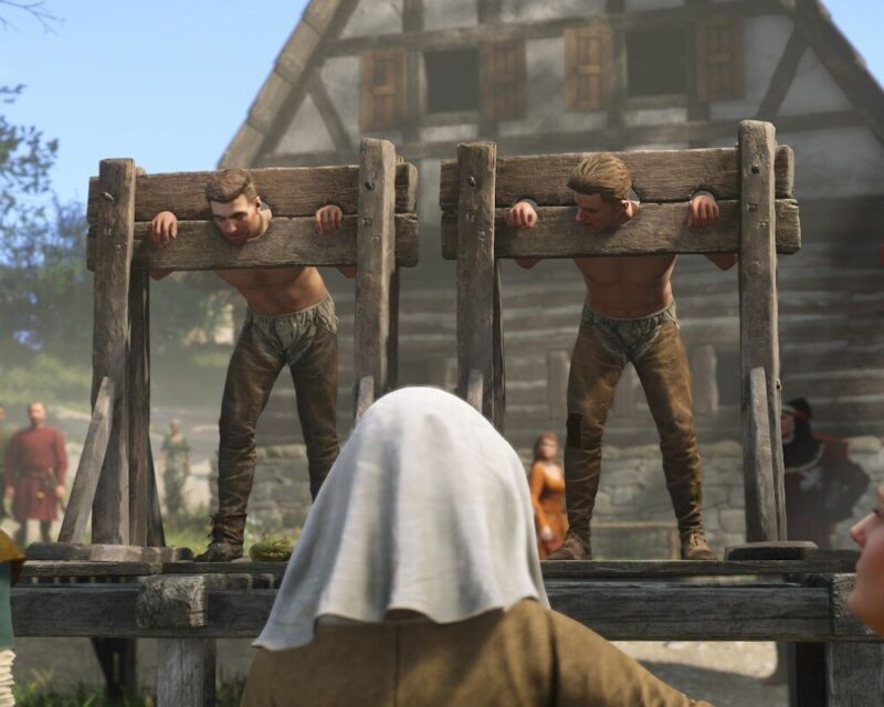 screenshot z gry Kingdom Come: Deliverance 2, widzimy na nim dwóch mężczyzn zakutych w dyby na podwyższeniu, w tyłu średniowieczna chata, a przed nimi widoczny tył głowy osłoniętej białą chustą