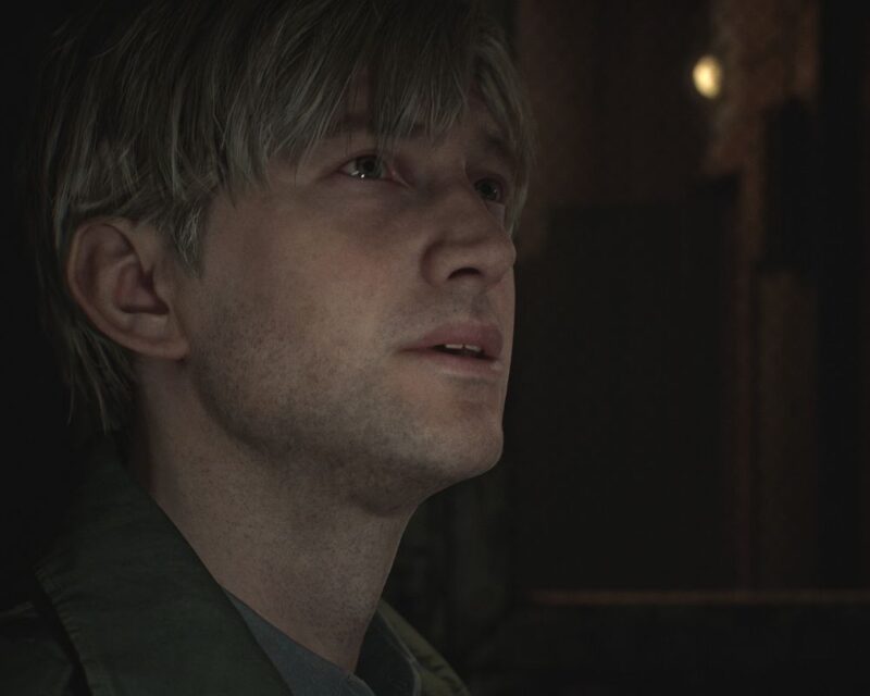 grafika z remake'a gry Silent Hill 2, przedstawia głównego bohatera, białego blondyna z lekkim zarostem, patrzy na coś nad sobą, w tle widać przyciemniony korytarz