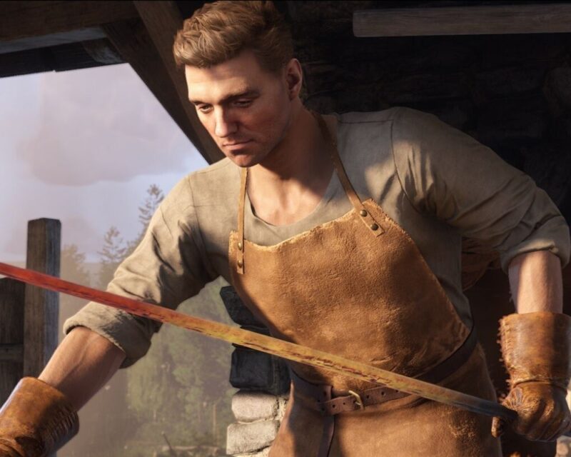 screenshot z gry Kingdom Come: Deliverance 2, widzimy mlodego mężczyznę o krótkich jasnych włosach, ubrany jest w ochronny fartuch kowalski, pochyla się nad bronią, którą własnie wykuwa