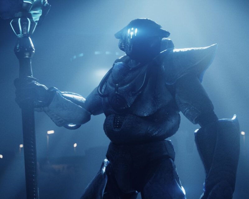 grafika z gry Helldivers 2 pokazująca nowego przeciwnika z aktualizacji Omens of Tyranny, jest ubrany w kombinezon bojowy, na głowie ma hełm ze świecącymi światełkami z przodu, w prawej ręce trzyma laskę, za nim widzimy zamazany pojazd