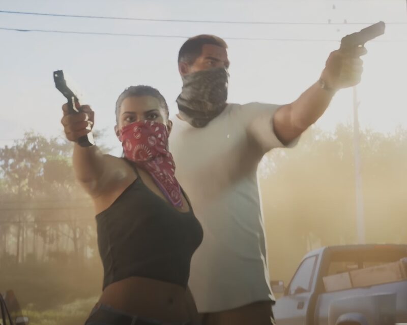 Na obrazku znajduje się protagonistka GTA 6. Jest ubrana w czarny top i ma czerwoną bandanę na ustach. Obok niej znajduje się mężczyzna w białej koszulce. Obydwoje celują z broni do wnętrza budynku. Bohaterowie są wyciemnieni, a za nimi znajduje się jasny krajobraz z drzewami.