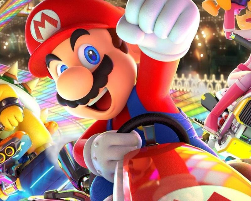 Na obrazku widać postać o dużych, niebieskich oczach, okrągłym nosie i czarnym wąsie. Ma na sobie czerwoną czapkę z białym kółkiem, w środku którego znajduje się duża czerwona litera „M”. Jest ubrana w czerwono-niebieski strój – czerwony podkoszulek i niebieskie ogrodniczki z żółtymi guzikami. Postać prowadzi mały czerwony pojazd przypominający gokart. W tle znajduje się kolorowa, tęczowa trasa, która lśni różnymi barwami. Widać też inne postacie – po lewej dużą, żółtą postać z kolczastą skorupą na plecach, a po prawej różową postać na motocyklu.