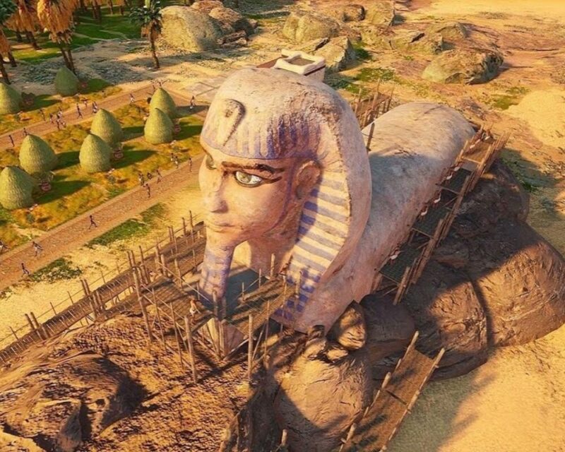 Kadr z gry *Builders of Egypt* przedstawiający monumentalną rzeźbę przypominającą Sfinksa w trakcie budowy, z widocznymi rusztowaniami i otaczającym ją krajobrazem Egiptu.