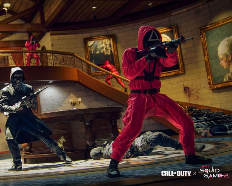 Grupa strażników w czerwonych uniformach z czarnymi maskami w kształcie geometrycznych symboli (trójkąt i kwadrat) stoi w eleganckim wnętrzu z drewnianymi balustradami i obrazami na ścianach. Jeden strażnik celuje z karabinu, podczas gdy w tle widać innych strażników patrolujących przestrzeń. W prawym dolnym rogu znajduje się logo wydarzenia „Call of Duty x Netflix Squid Game 2”.