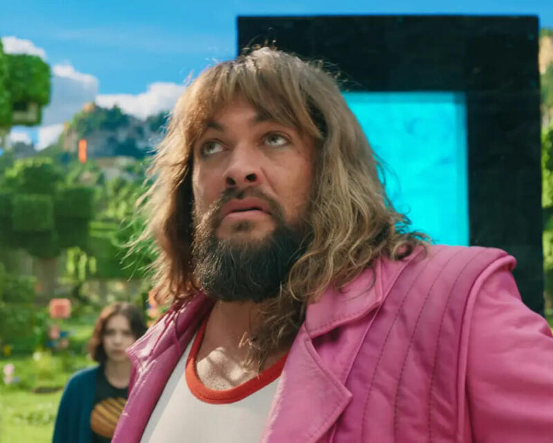 Aktor Jason Momoa, ubrany w różową kurtkę, stoi na tle krajobrazu z krainy Minecraft. W tle widoczne są pikselowe drzewa, zielone wzgórza i charakterystyczny portal Nether z ciemnych bloków.