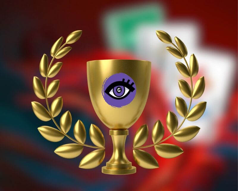 tło w większości ciemnoczerwone, w górnym prawym rogu widać rozmyty ksztalt kart, na pierwszym planie złoty laur zwycięzcy z pucharem na srodku, na pucharze fioletowe logo portalu Grajmerki w formie kobiecego oka na fioletowym tle