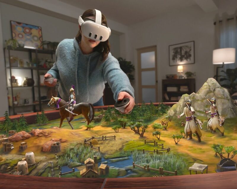 Na obrazku widzimy kobietę, która ma na sobie gogle VR i trzyma w rękach kontrolery. Kobieta stoi przed stołem, na którym znajduje się makieta przedstawiająca bitwę. Makieta zawiera miniaturowe figurki żołnierzy, zwierząt i budynków. Kobieta wydaje się wchodzić w interakcję z makietą za pomocą gogli i kontrolerów.