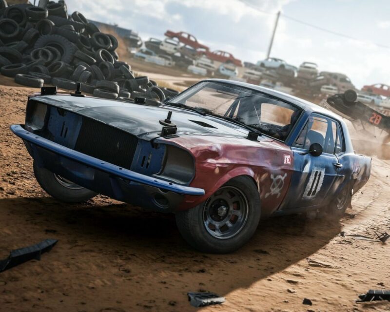 Na zdjęciu widać stary, mocno przerobiony samochód na piaszczystym torze wyścigowym. Auto ma zniszczoną karoserię i jest pomalowane na niebiesko-czerwono-czarno. W tle widać stos opon i wraki innych samochodów. Jeden pojazd właśnie koziołkuje w powietrzu. Wokół unosi się kurz. Wreckfest 2 data premiery wczesny dostęp