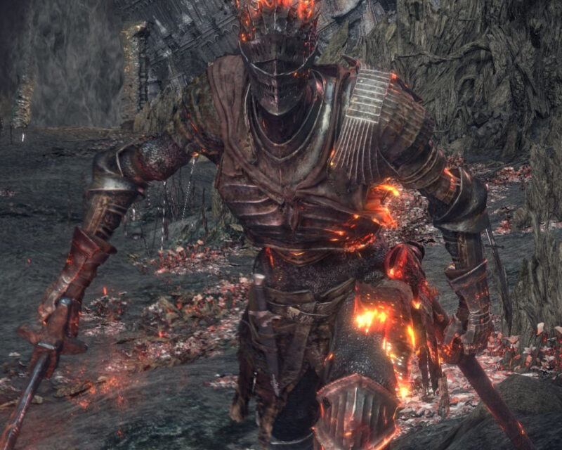 Na obrazku znajduje się boss z gry Dark Souls 3, czyli Soul of Cinder. Ma ciemną zbroję i częściowo się pali. Obrazek jest bardzo ciemny. Postać jest gotowa do walki.
