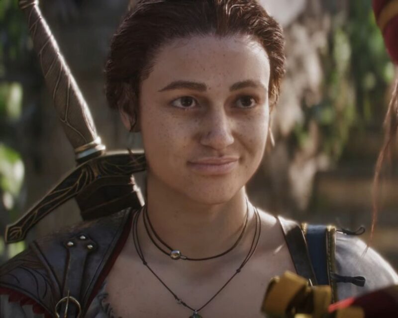 Postać z gry „Fable 4” o krótkich, brązowych włosach i piegach na twarzy, uśmiecha się delikatnie. Ubrana jest w zbroję i naszyjniki, a na plecach nosi miecz o zdobionej rękojeści. W tle widać roślinność i kamienne schody, co nadaje scenie przygodowego klimatu.