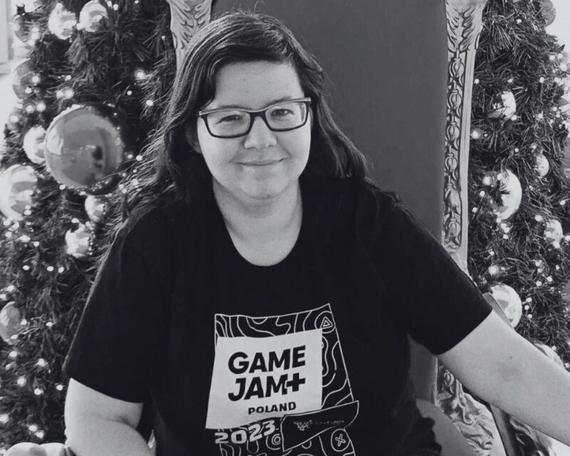 czarno białe zdjęcie naszej redaktorki, Izy, w bluzce z napisem Game Jam i z uśmiechem na twarzy - Iza, bardzo Cię nam brakuje