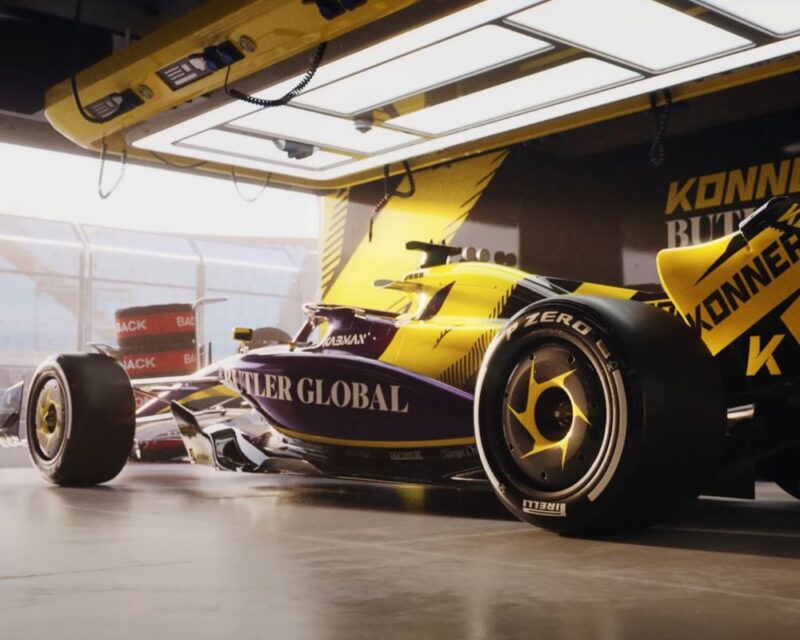 Na obrazku jest bolid wyścigowy, który stoi w grażu stoi w garażu, oświetlony od góry. Na jego nadwoziu widnieją logotypy sponsorów. W tle widać barierki toru wyścigowego i stos opon.