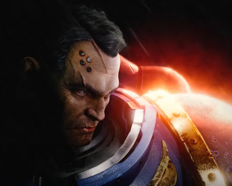 Na obrazku znajduje się bohater z gry Warhammer 40k Space Marine 3. Ma skupiony wyraz twarzy i ciemne włosy. W twarzy ma metalowe elementy. Jest ubrany w masywną, granatową zbroję. Za nim znajduje się błysk. Całość jest bardzo ciemna.